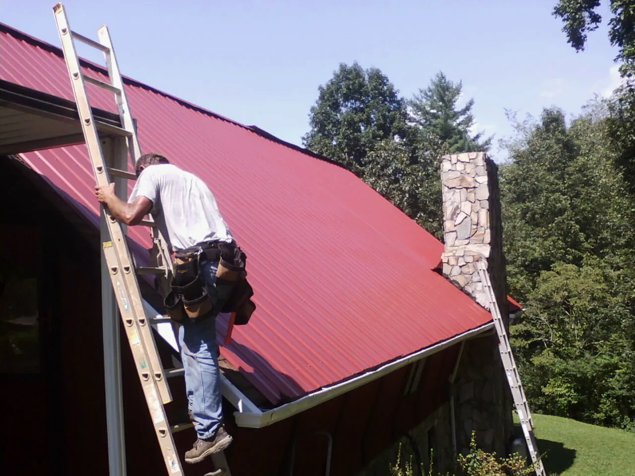 Roofer on a ladder beside a red metal roof and stone chimney — Soffit & Fascia Repair by Roofing Brothers in Prospect